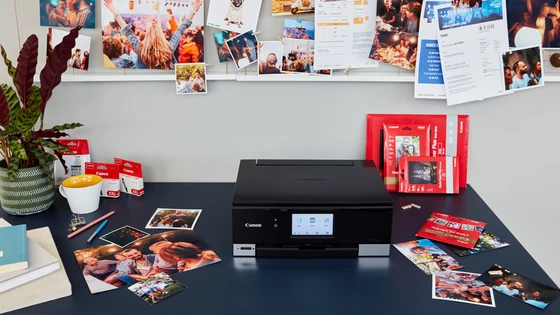 Принтер стоит на столе, вокруг него лежат фотобумага, изображения и упаковки чернил, а над принтером висит доска с различными отпечатками.