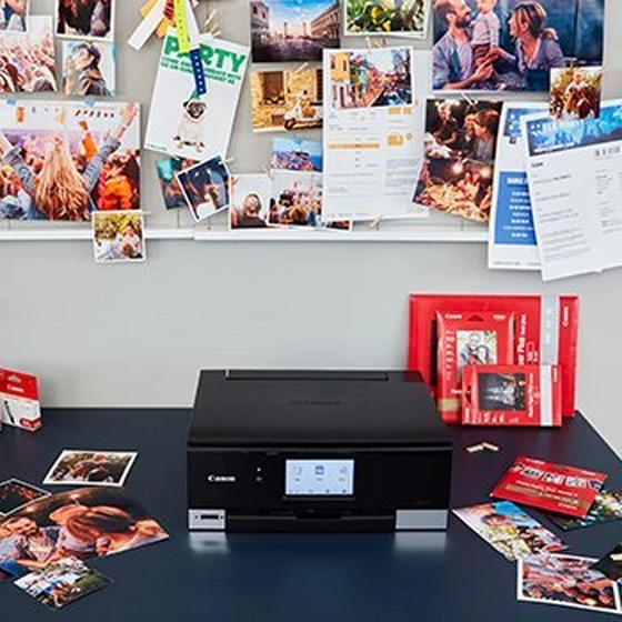 A desk with a printer, photo paper, images and ink packages around it and a board with different printed items above it.