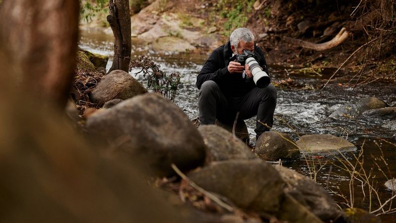 A white-haired man crouches on the edge of a stream, pointing a Canon camera with a long white lens at the flowing water.