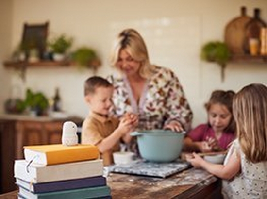 A woman and her three children standing around a mixing bowl in a kitchen as they prepare to bake something. The PowerShot PX sits on top of a pile of books facing them.