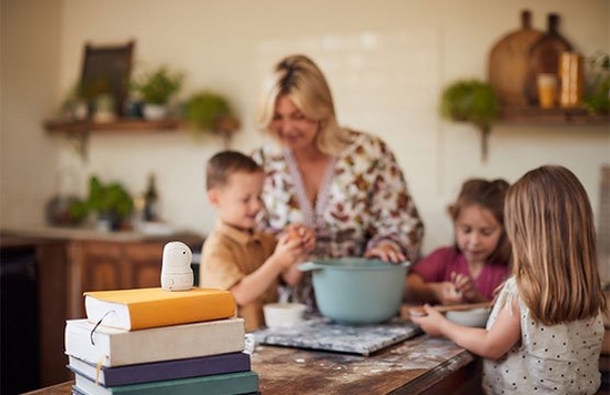 A woman and her three children standing around a mixing bowl in a kitchen as they prepare to bake something. The PowerShot PX sits on top of a pile of books facing them.