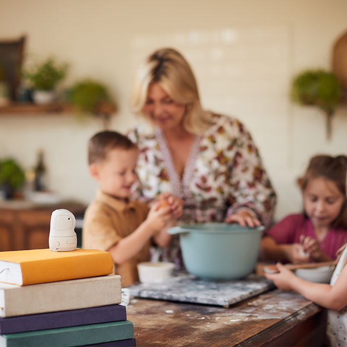 A woman and her three children standing around a mixing bowl in a kitchen as they prepare to bake something. The PowerShot PX sits on top of a pile of books facing them.