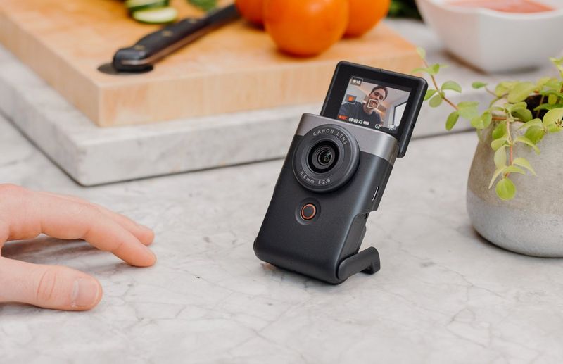 A Canon PowerShot V10 stands on a kitchen worktop with a chopping board in the background and a vlogger's hand resting on the surface in front of it.