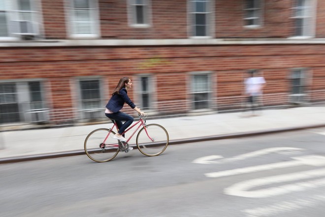 Um ciclista na rua de uma cidade com um edifício de tijolos e um peão com fundo desfocado devido ao efeito de "panning".