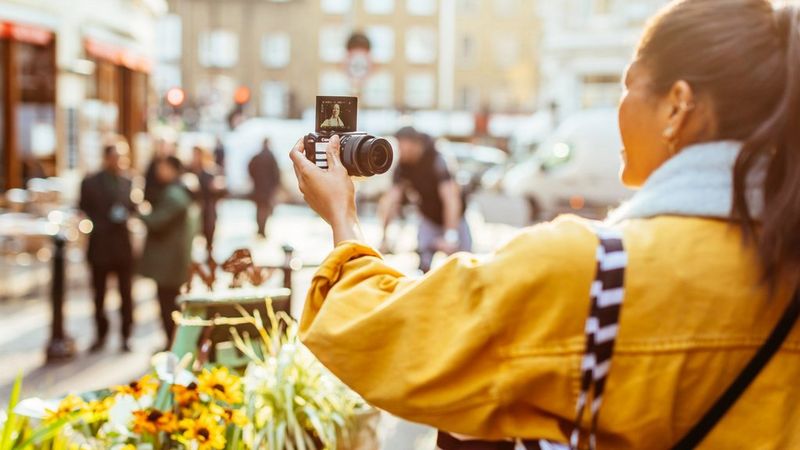 A young woman wearing a yellow jacket takes a selfie with the Canon M50 in a city environment.