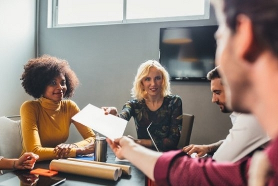 Four smiling people sitting around a desk. A woman passing a man a piece of paper.