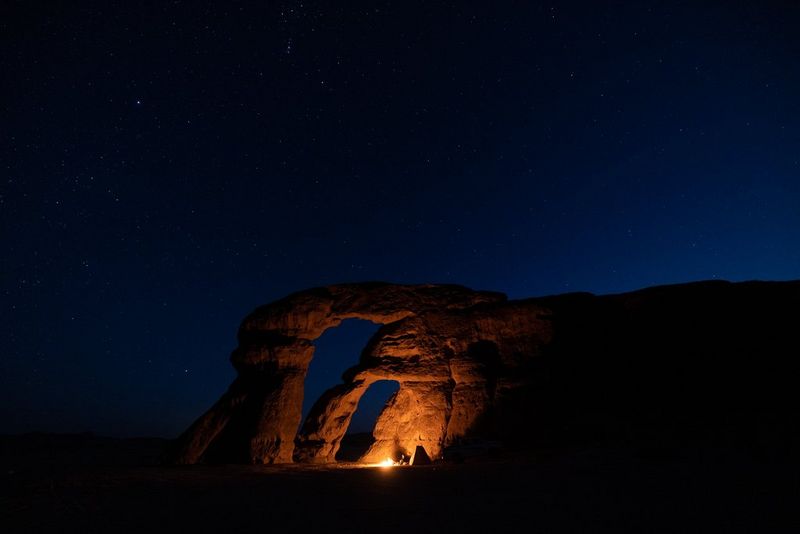 A small fire glows in a rocky cave, while the night sky is filled with stars above.