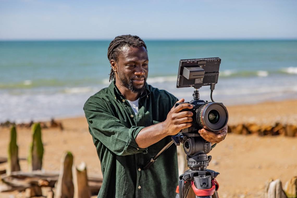 Against a serene beach backdrop, filmmaker Jolade Olusanya smiles as he studies his footage, filmed on the Canon EOS C70.