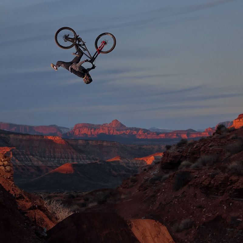 A mountain bike with rider is upside down, travelling through the air above a rocky desert landscape.