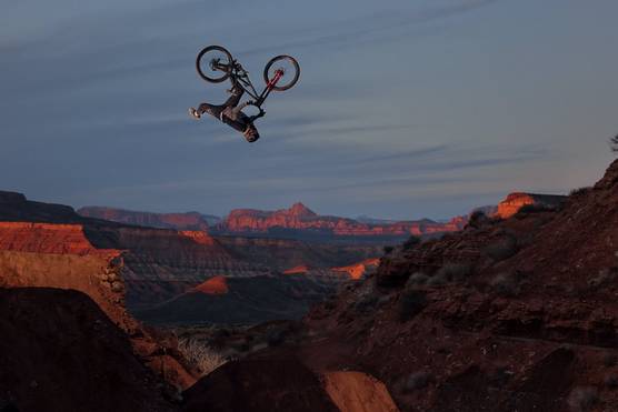 A mountain bike with rider is upside down, travelling through the air above a rocky desert landscape.