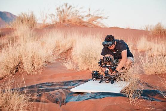 In a field, a man wearing goggles on his forehead crouches next to a Canon EOS R5 C attached to a drone.