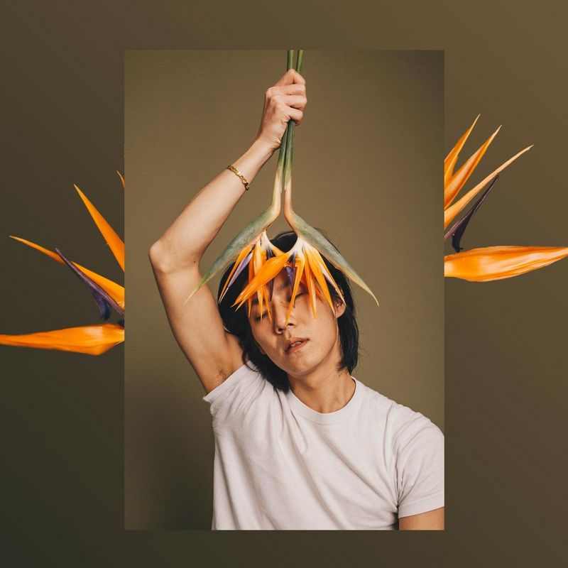 Un homme vêtu d'un t-shirt blanc tient des fleurs orange devant son visage devant un fond marron. Cette image est en surimpression sur une autre photo des mêmes fleurs.