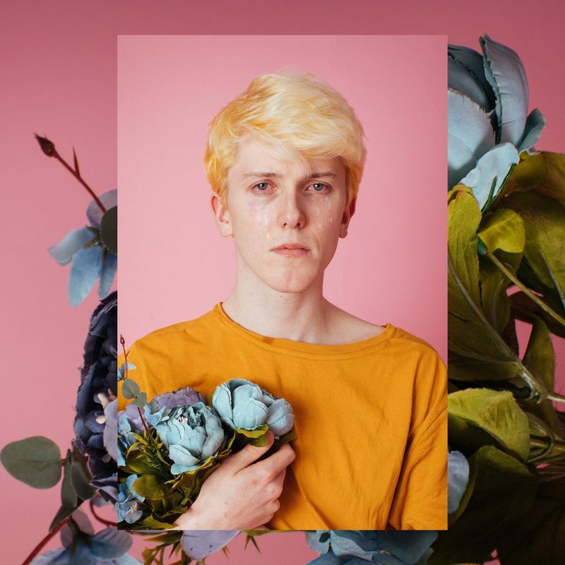 Un homme aux cheveux blonds décolorés tenant des fleurs bleues et pleurant pose devant un fond rose. Cette image est en surimpression sur une autre photo des mêmes fleurs.