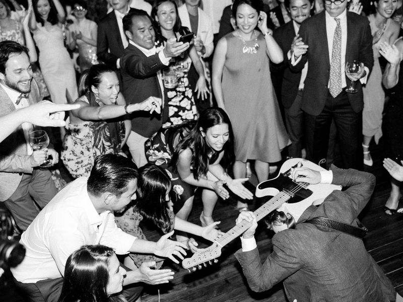 Image en noir et blanc d'un musicien s'agenouillant sur le sol avec sa guitare face à une foule d'invités qui l'acclament.