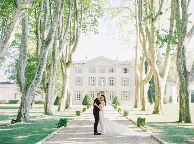 Photo en couleur de mariés dans une allée bordée d'arbres devant une maison de maître aux couleurs vert pâle et crème.