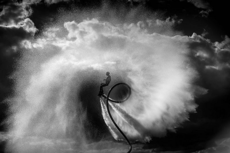 Une photographie en noir et blanc d'une personne faisant du flyboard sur un lac, des éclaboussures d'eau virevoltant tout autour d'elle.