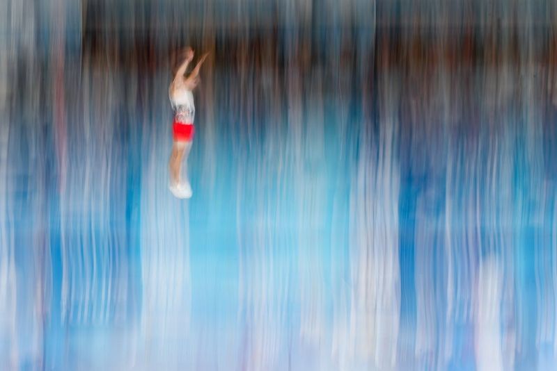 Un jeune trampoliniste portant un haut blanc et un short rouge est photographié en plein saut. Lui et l'arrière-plan sont flous.