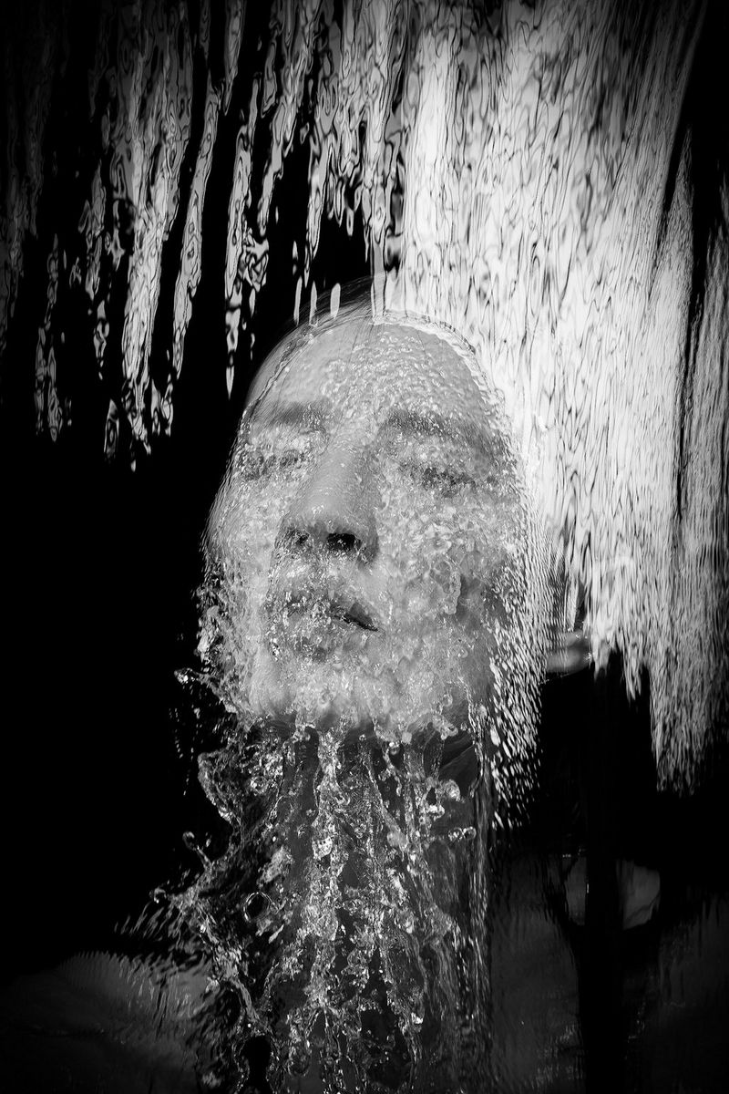 Une image en noir et blanc représentant de l'eau qui tombe en cascade sur le visage d'une femme incliné vers le haut.