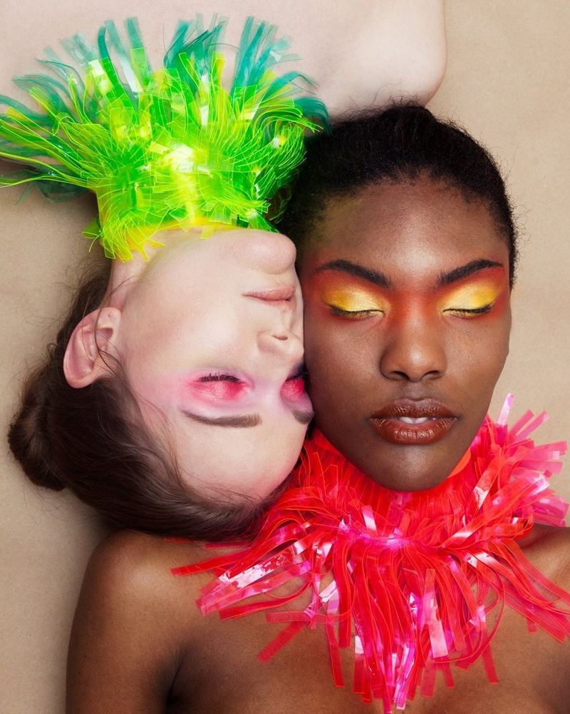 Deux mannequins allongées portant des fards à paupières colorés et des pièces de cou élaborées, la tête l'une contre l'autre et les yeux fermés.