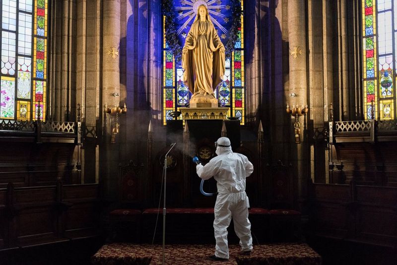 Une silhouette en combinaison de protection blanche vaporise une église richement décorée avec une statue de Marie et des vitraux.