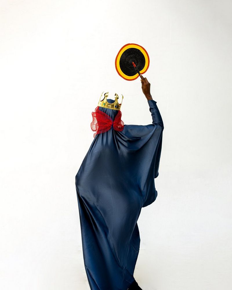 Une mannequin drapée dans un tissu bleu foncé satiné, un morceau de tulle rouge noué autour du cou et une couronne dorée sur la tête. De sa main levée, elle tient un éventail.