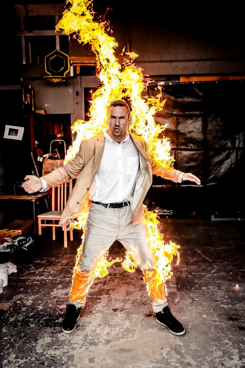 Un cascadeur en costume beige fait la grimace, son dos et ses jambes enveloppés par les flammes.