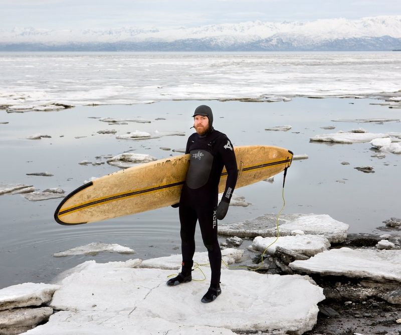 Un homme barbu en combinaison tient une planche de surf sous son bras. Il est debout à côté d'une étendue d'eau glaciale.
