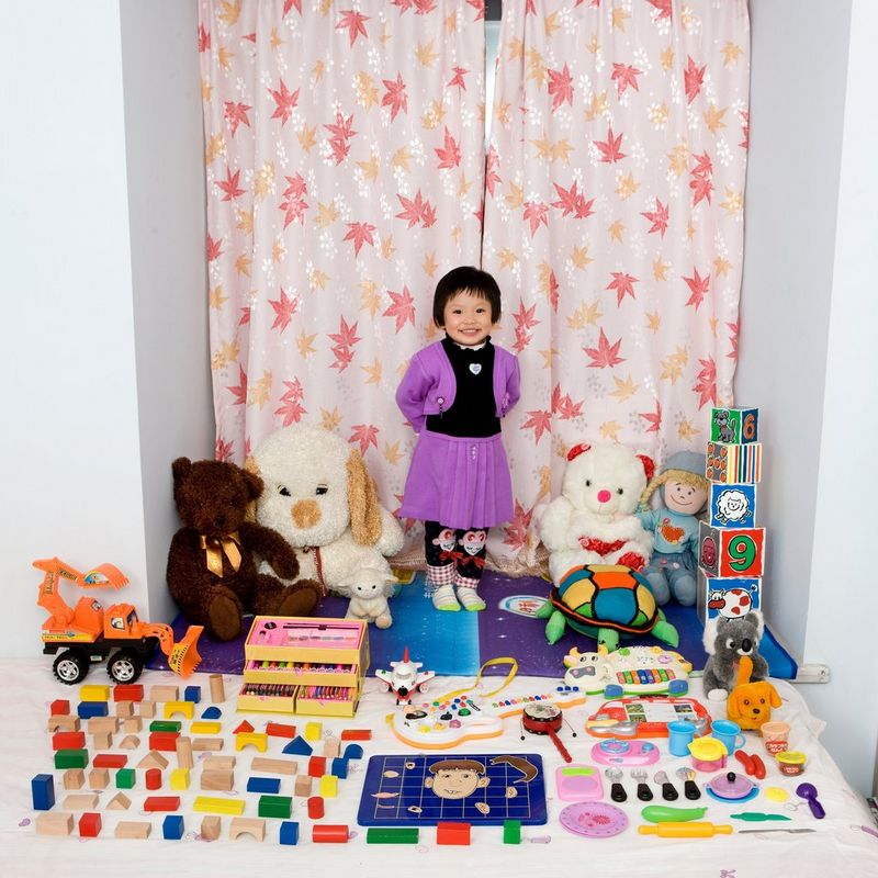 Une petite fille de trois ans sourit fièrement en montrant ses jouets préférés, notamment deux gros ours en peluche.