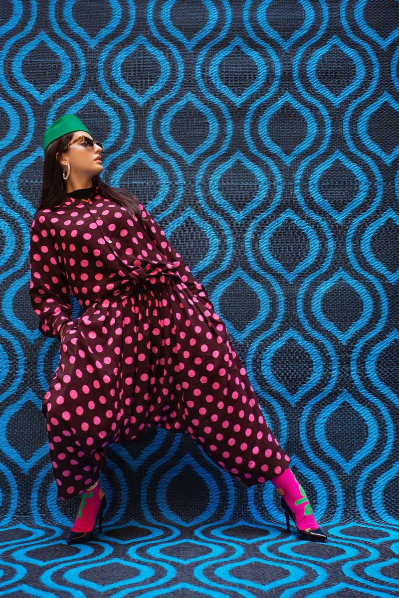 Une jeune femme portant un chapeau vert et une combinaison noire à pois roses pose devant un fond bleu aux motifs saisissants.