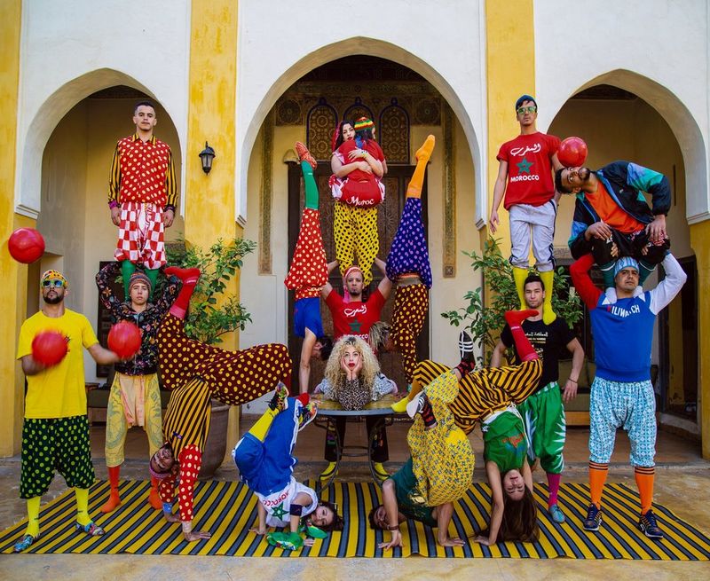 Un groupe d'hommes et de femmes vêtus de vêtements aux couleurs vives effectue une pose acrobatique devant un bâtiment blanc avec des piliers jaunes.