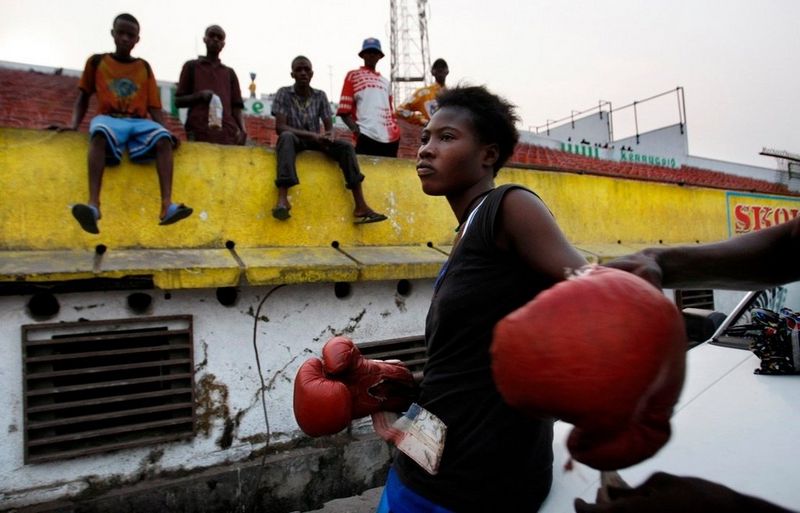 Un entraîneur aide une jeune femme à enfiler des gants de boxe, pendant qu'un groupe d'hommes observent la scène.