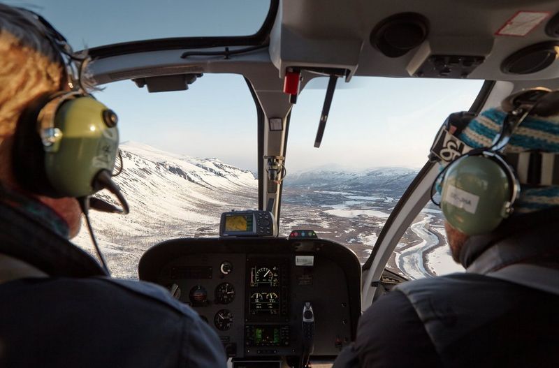 Deux hommes portant des casques d'aviation observent un paysage enneigé depuis le cockpit d'un hélicoptère.
