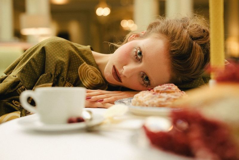 Une jeune femme portant de longs faux cils sur la ligne des cils inférieurs pose sa tête sur une table à côté d'un gâteau et d'une tasse avec soucoupe.