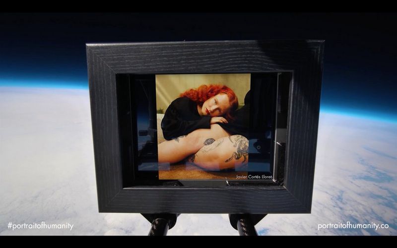 Image d'une femme aux cheveux orange vif, posant sa tête sur les jambes tatouées d'une autre femme, exposée dans l'espace.