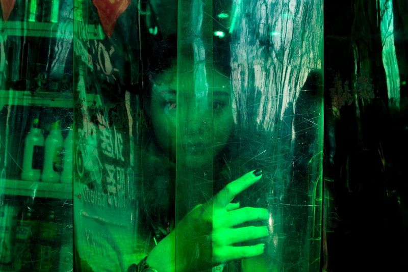 Un cliché, teinté de vert, d'une jeune streameuse regardant à l'extérieur dans l'encadrement de la porte, derrière une bâche transparente.