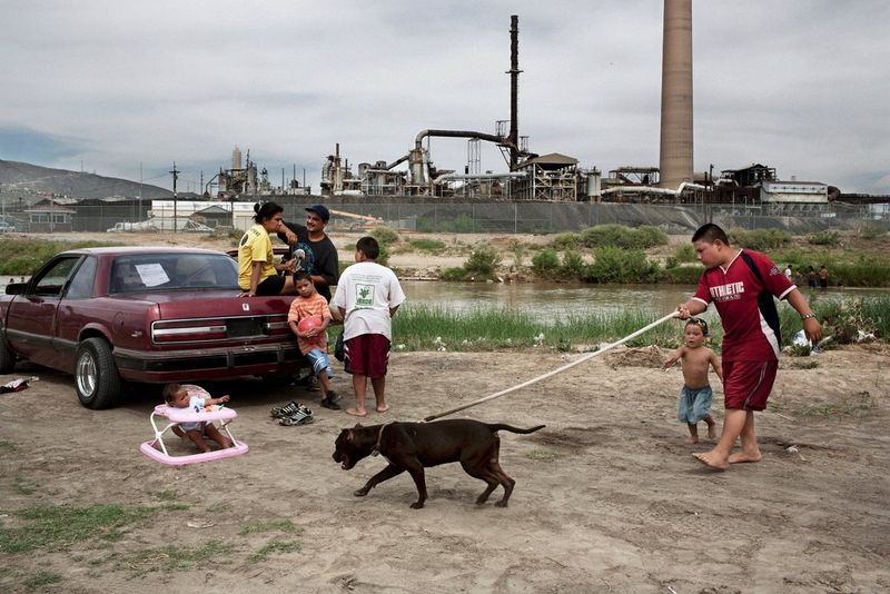 À la frontière entre les États-Unis et le Mexique, une femme est assise sur l'arrière de sa voiture, tandis que son mari et ses cinq enfants jouent et discutent autour d'elle. Derrière eux, on peut voir la fonderie de l'usine ASARCO à El Paso, au Texas.