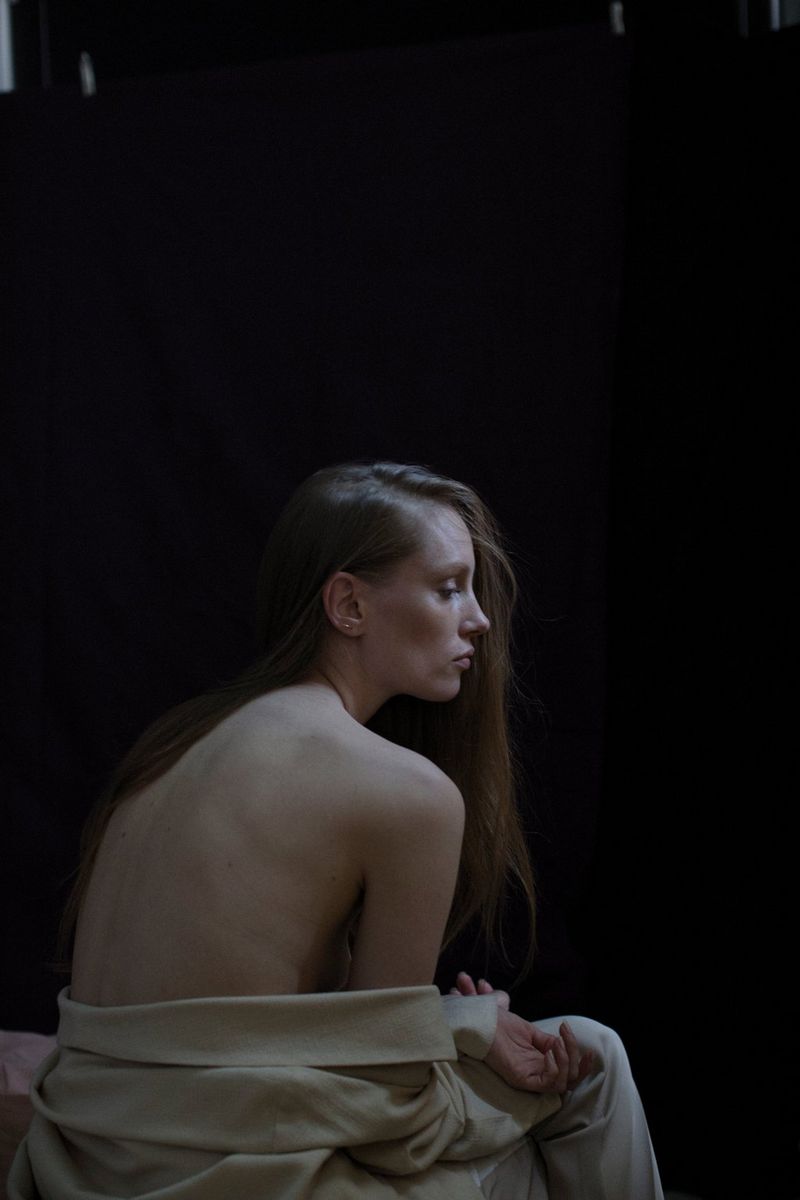 Une jeune femme rousse, dont le haut du corps est dénudé, est assise dos à l'objectif.