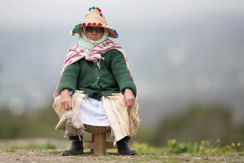 Une femme âgée, vêtue de vêtements traditionnels marocains et tenant des sacs de céréales, est assise sur un petit tabouret en bois au bord de la route.