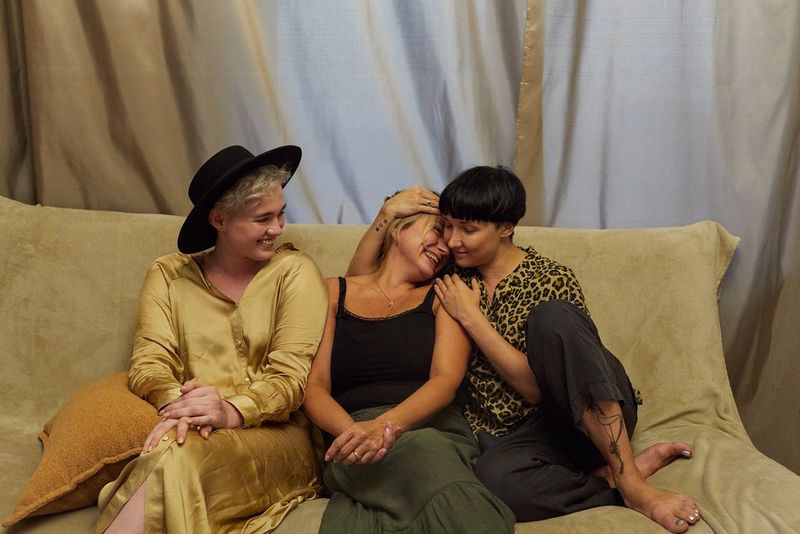 Une mère et sa fille adulte s'étreignent sur un canapé. À côté d'elles, une femme portant un chapeau noir sourit.