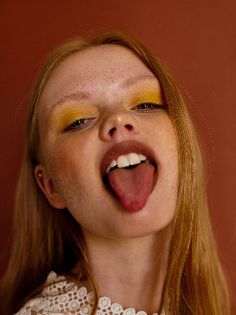 Une jeune mannequin aux longs cheveux roux et portant du fard à paupières jaune tire la langue à l'objectif.
