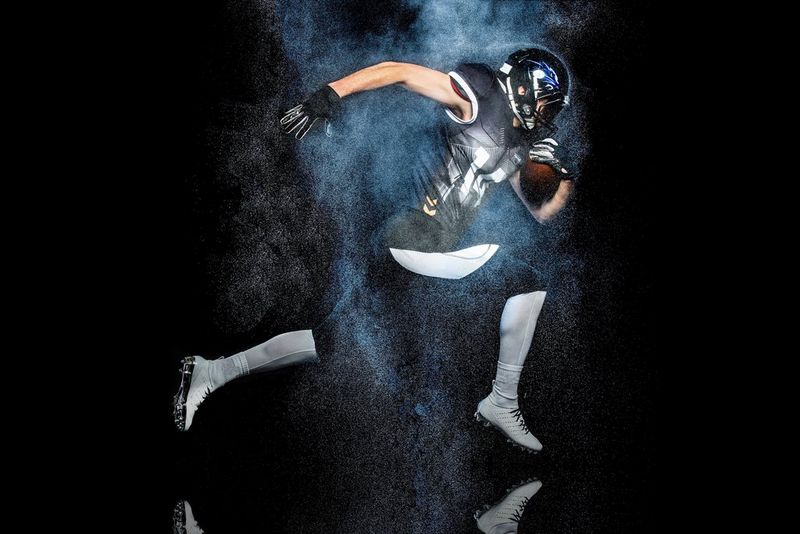 Un joueur de football américain pose à mi-course, serrant un ballon devant lui avec un bras et une jambe tendus vers l'arrière, sur un fond noir.