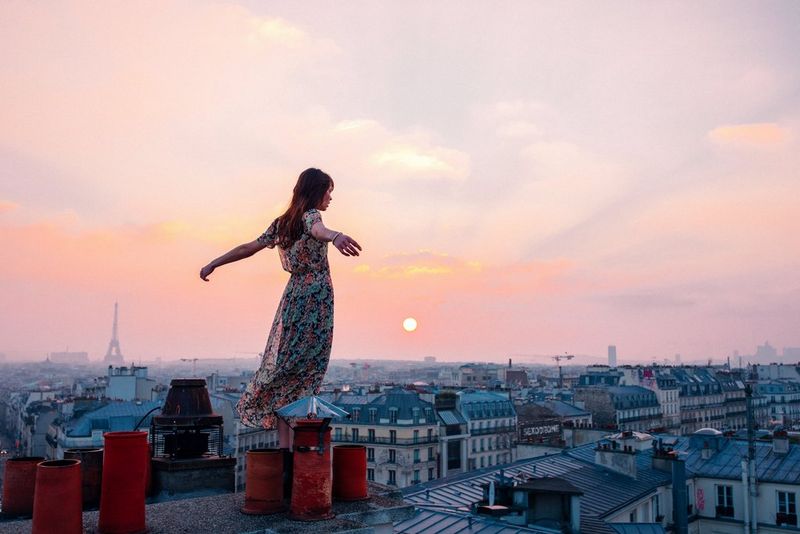 Femme portant une longue robe flottante debout sur un toit surplombant les rues de Paris. Ses bras sont étirés derrière elle.