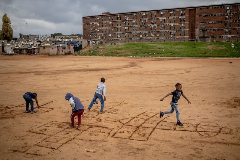 Alt text : Quatre jeunes enfants jouent à la marelle sur un lot de terre poussiéreux devant un grand bâtiment.