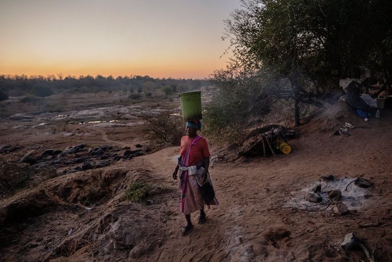 Une femme sud-africaine portant un grand seau rempli d'eau de rivière sur sa tête traverse un paysage boueux et dépouillé.