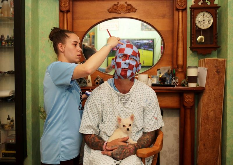 Un fan du club Burnley FC tient dans ses bras un petit chien alors que sa fille lui peint le visage avec des carreaux bordeaux et bleus avant de regarder un match à la télévision pendant le confinement.