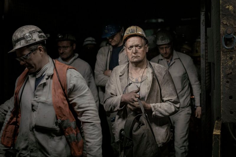 German miners covered in coal dust and wearing hard hats emerge from a narrow, dark elevator. 