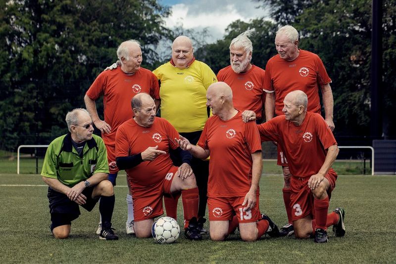 Un groupe de footballeurs seniors en tenues rouges discutent amicalement tandis qu'ils se préparent à poser pour leur photo d'équipe.