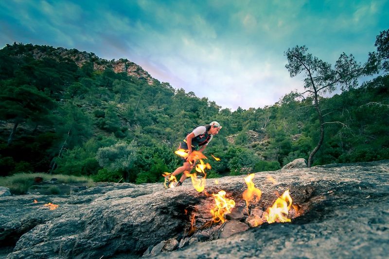 Un coureur d'ultra-marathon portant une casquette court à travers un paysage rocheux. On aperçoit un petit feu au premier plan.