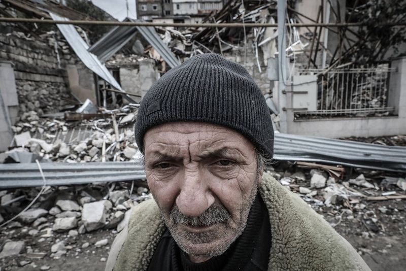 Un vieil homme portant un bonnet en laine se tient devant les décombres d'une maison détruite.