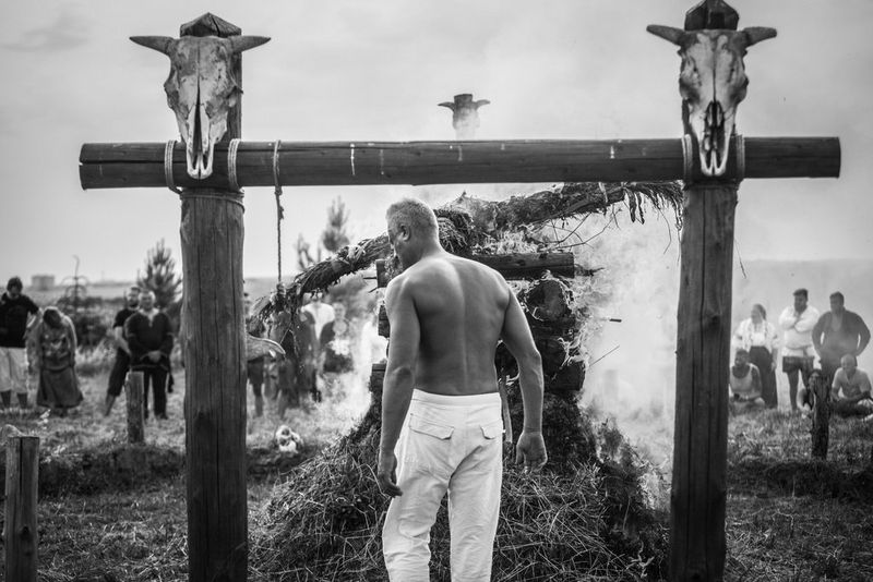 Un homme torse nu debout entre deux poteaux ornés de crânes d'animaux. Une effigie brûle dans un bûcher devant lui.